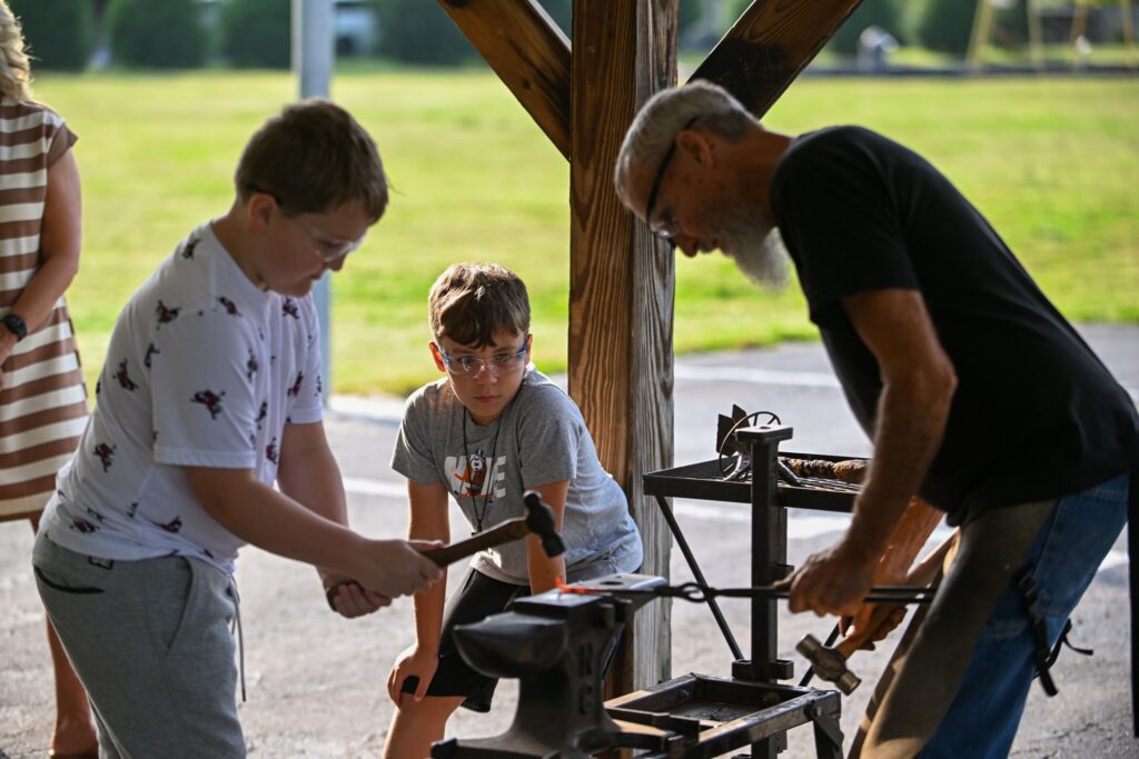 Two young students and a blacksmith forge metal on an anvil