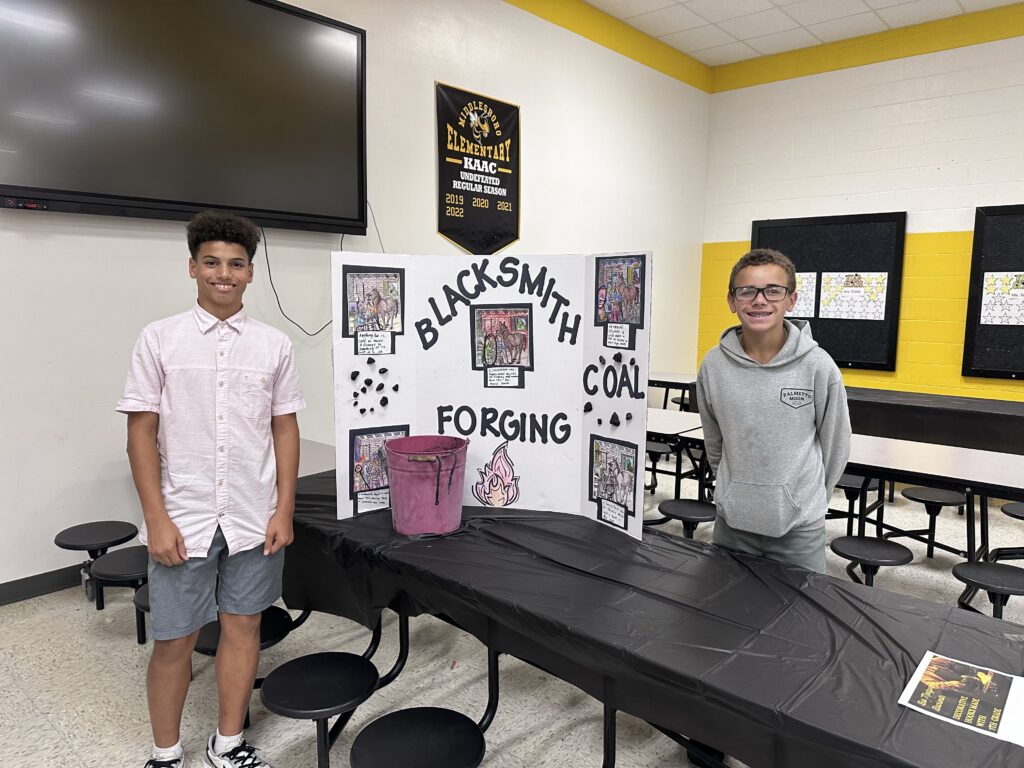 Two young students with a project board display about blacksmithing